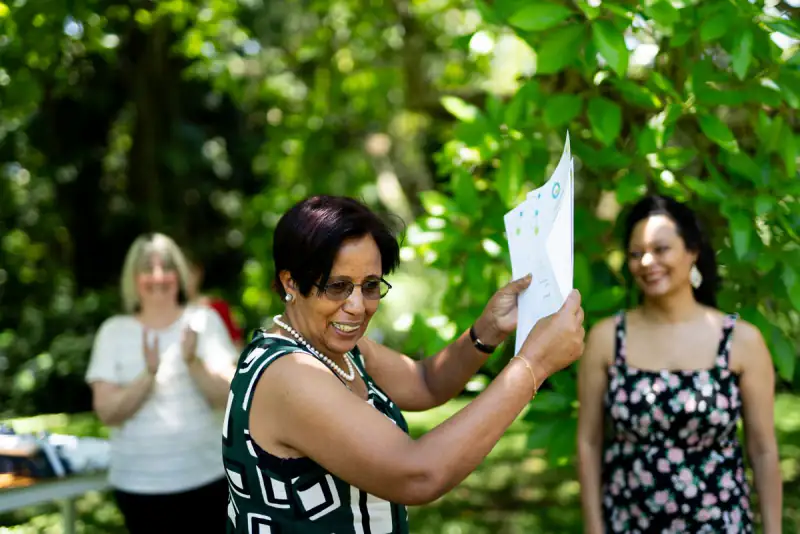 A course participant smiles as she holds up her certificate in a garden