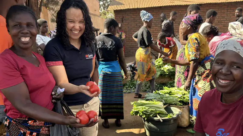 A Sense consultant and women market traders smiling with fresh tomatoes and vegetables