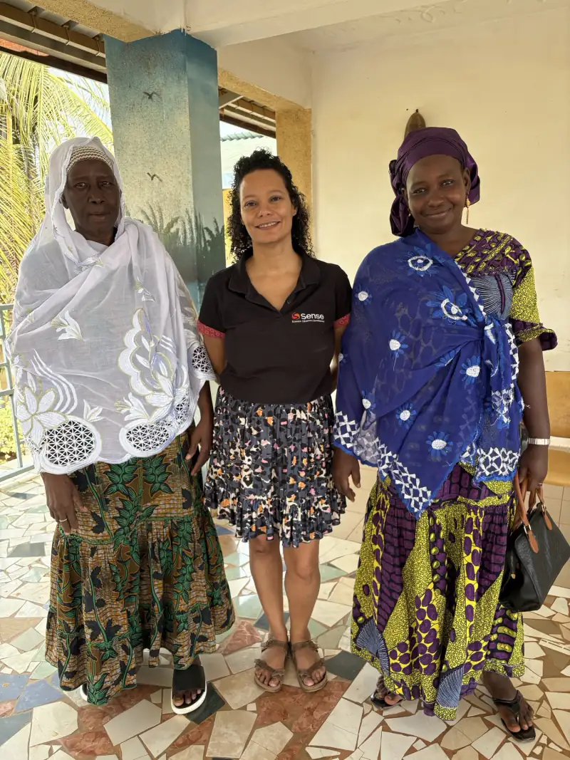 Three women standing together and smiling on a tiled veranda, one in a Sense shirt